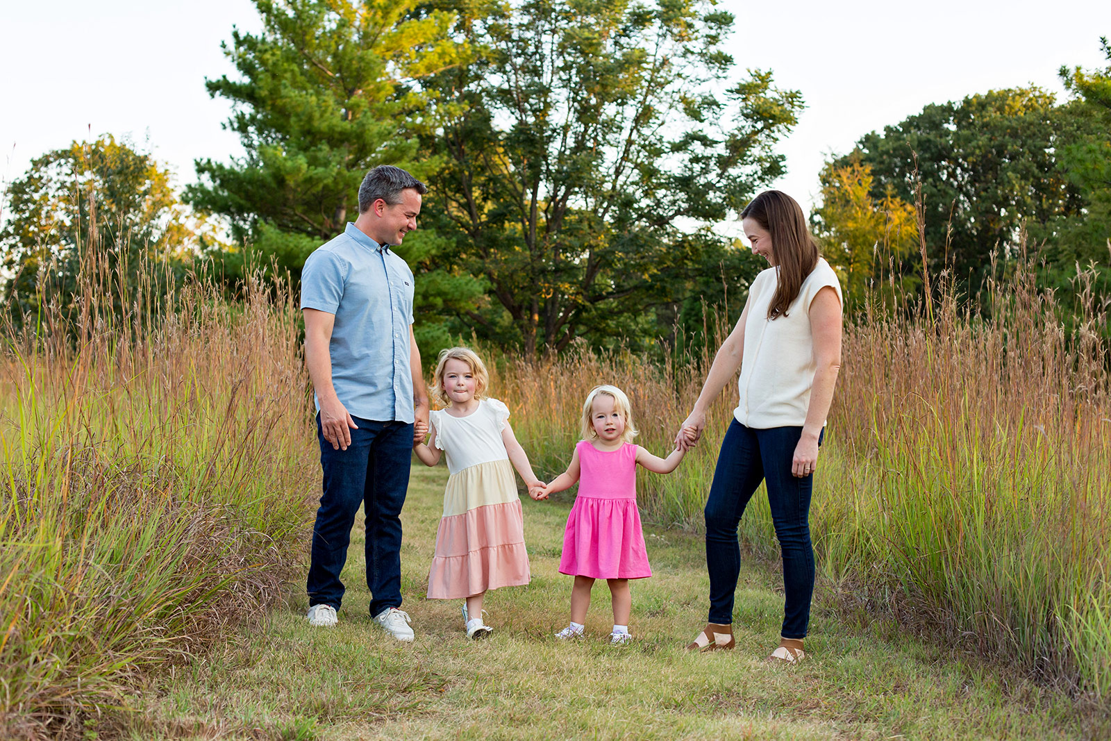 John and Ella hold hands with their girls as they walk through tall grasses.