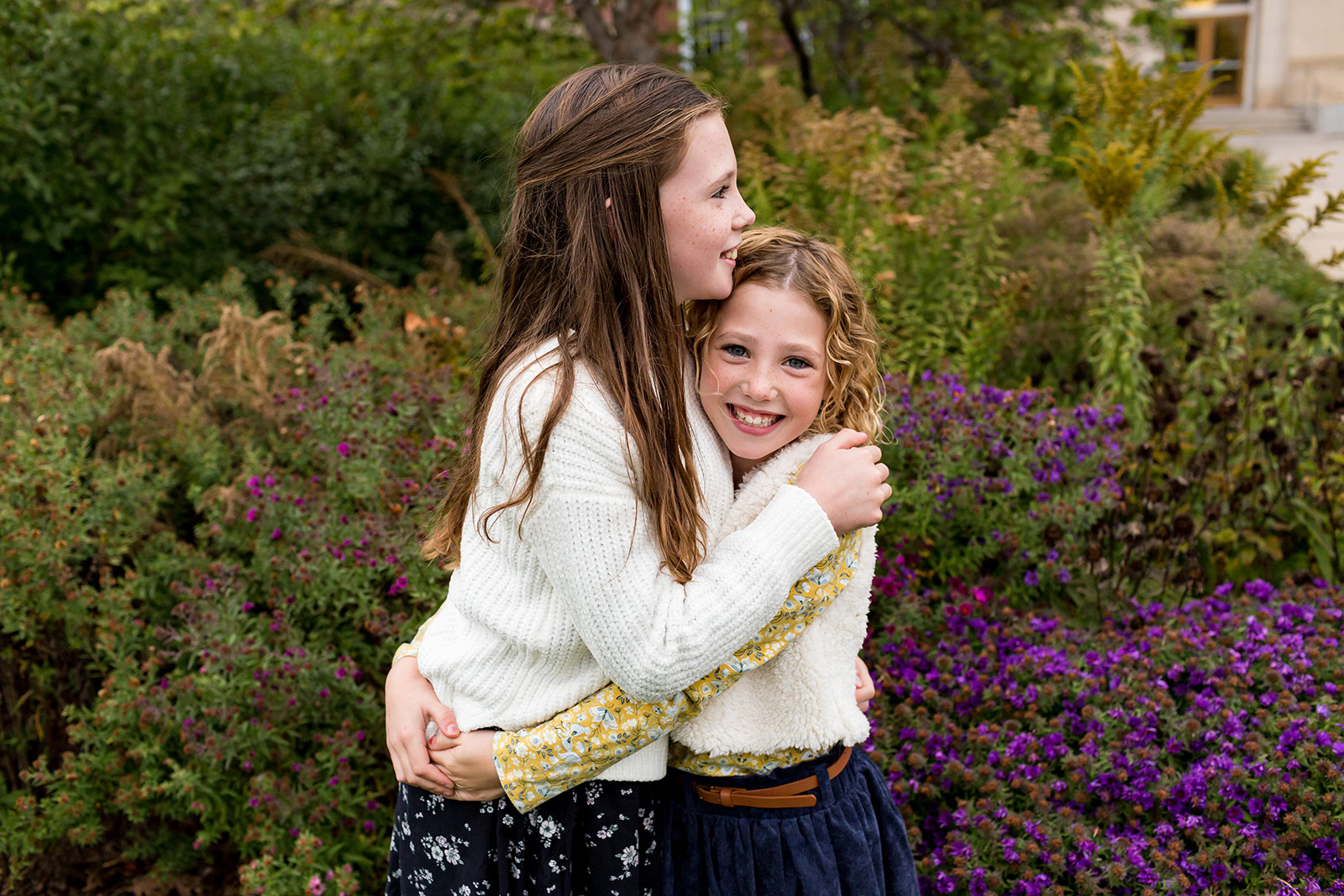 Nora and Harper hug each other tight in front of purple flowers.