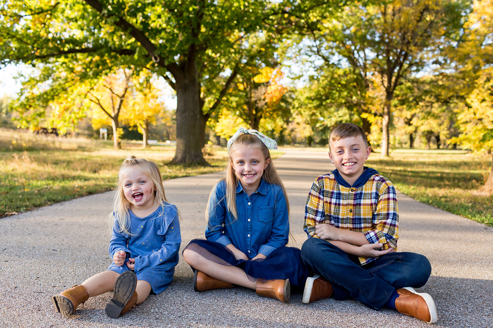 All three kids sit on the path in front of fall trees.