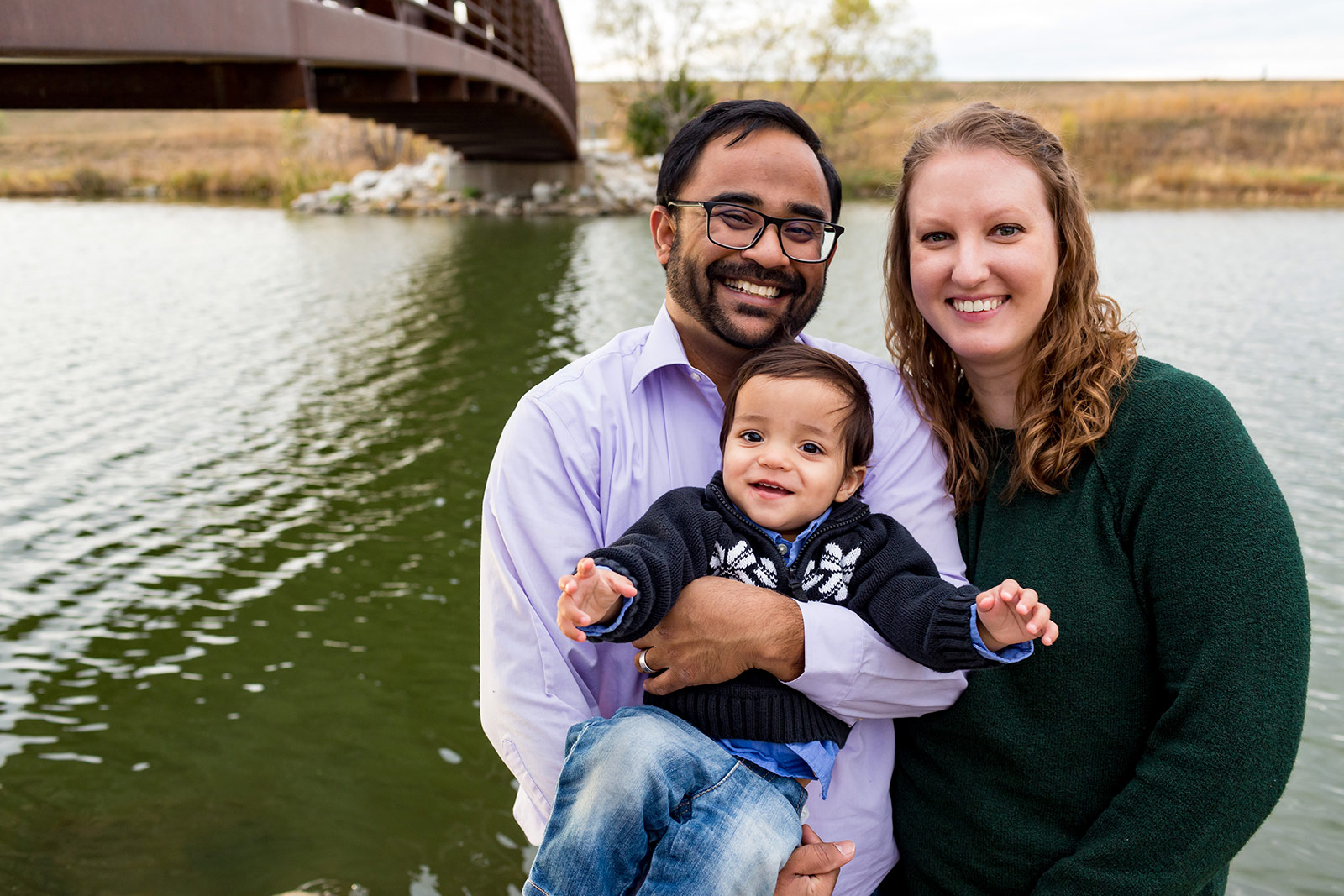 The family smiles naturally for a portrait in front of a lake and bridge.