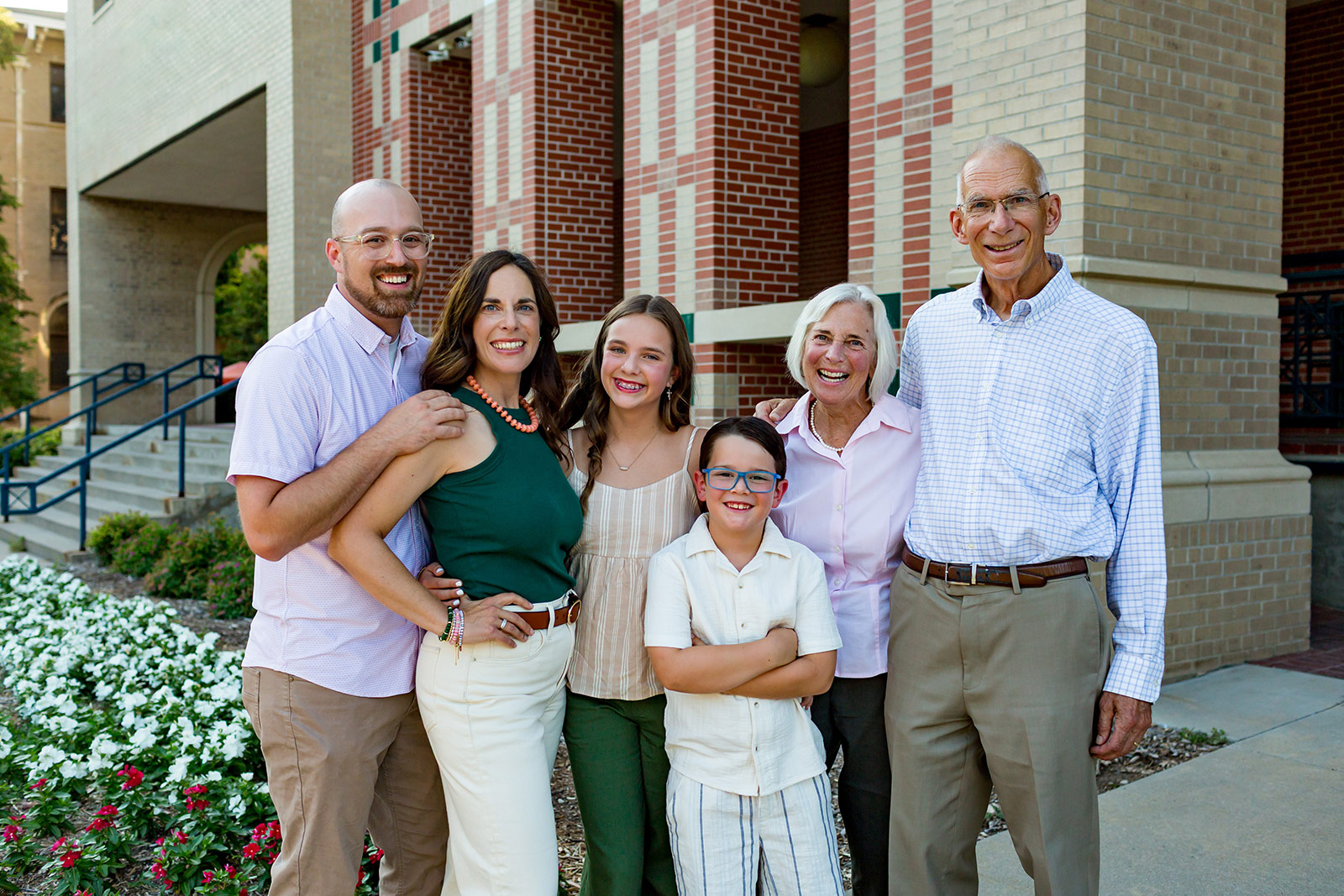 Three generations of a family pose outside a brick campus building, with two grandparents, two parents, and two children smiling at the camera.