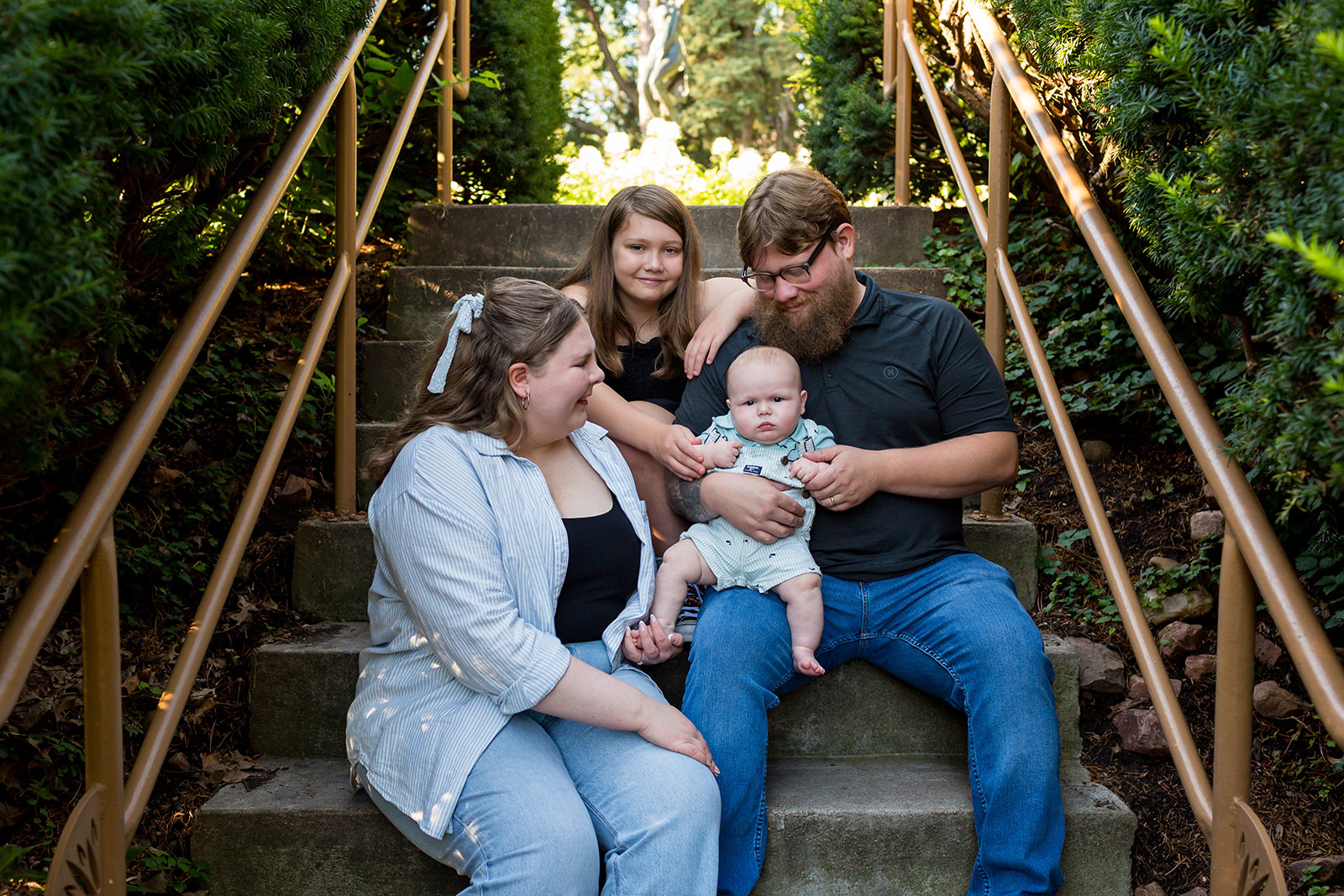Classic family portrait on garden steps with parents, their baby, and older daughter sharing a relaxed moment that highlights genuine emotion, natural connection, and authentic family photography.