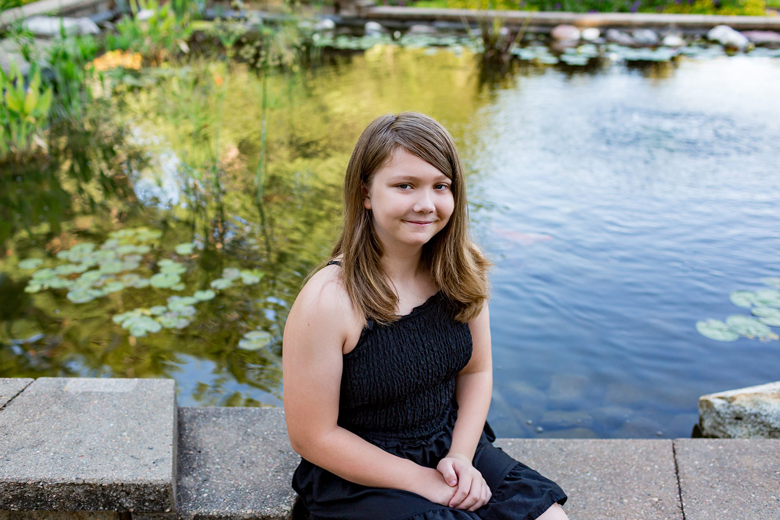 Natural child portrait of a young girl sitting beside a peaceful garden pond, smiling softly during a relaxed outdoor family photography session that captures her personality and genuine expression.