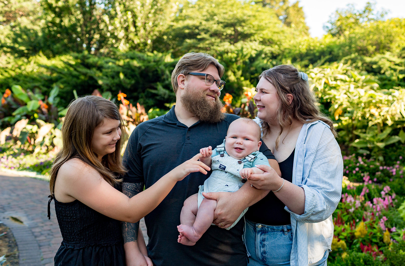 Parents and older sister smiling at a happy baby while standing together among the flowers at Sunken Gardens in Lincoln, Nebraska.