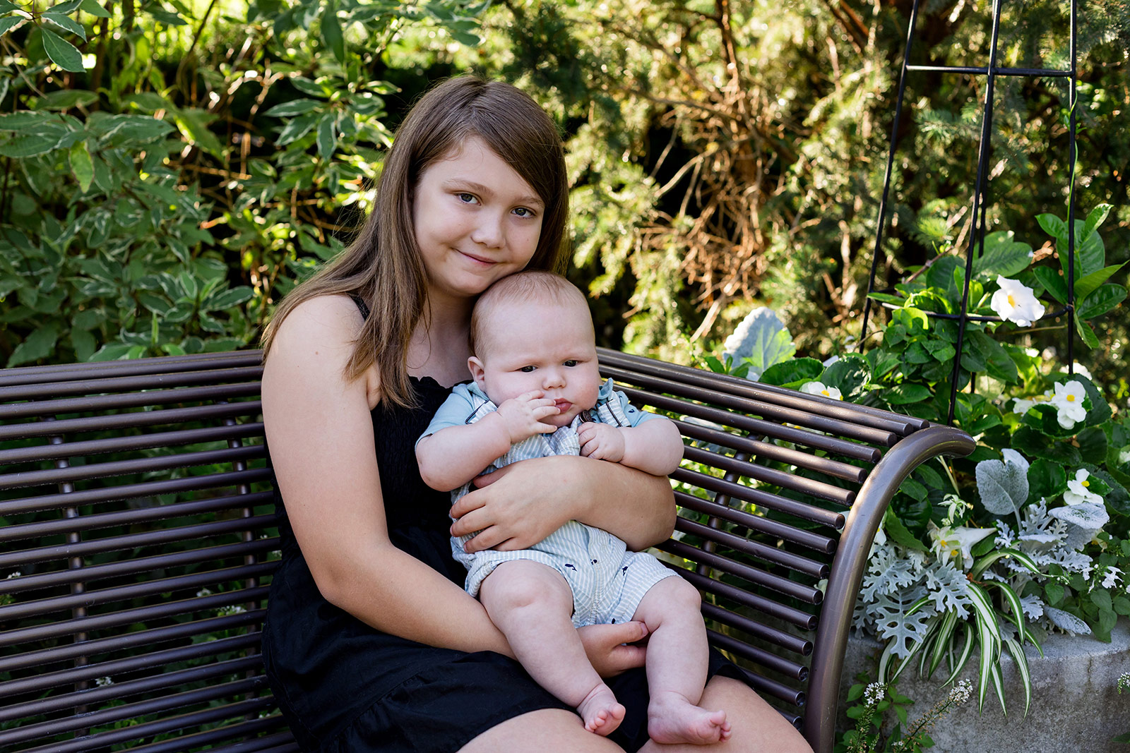 Older sister holding her baby brother on a garden bench, creating a sweet sibling portrait during a family photography session in Lincoln, Nebraska.