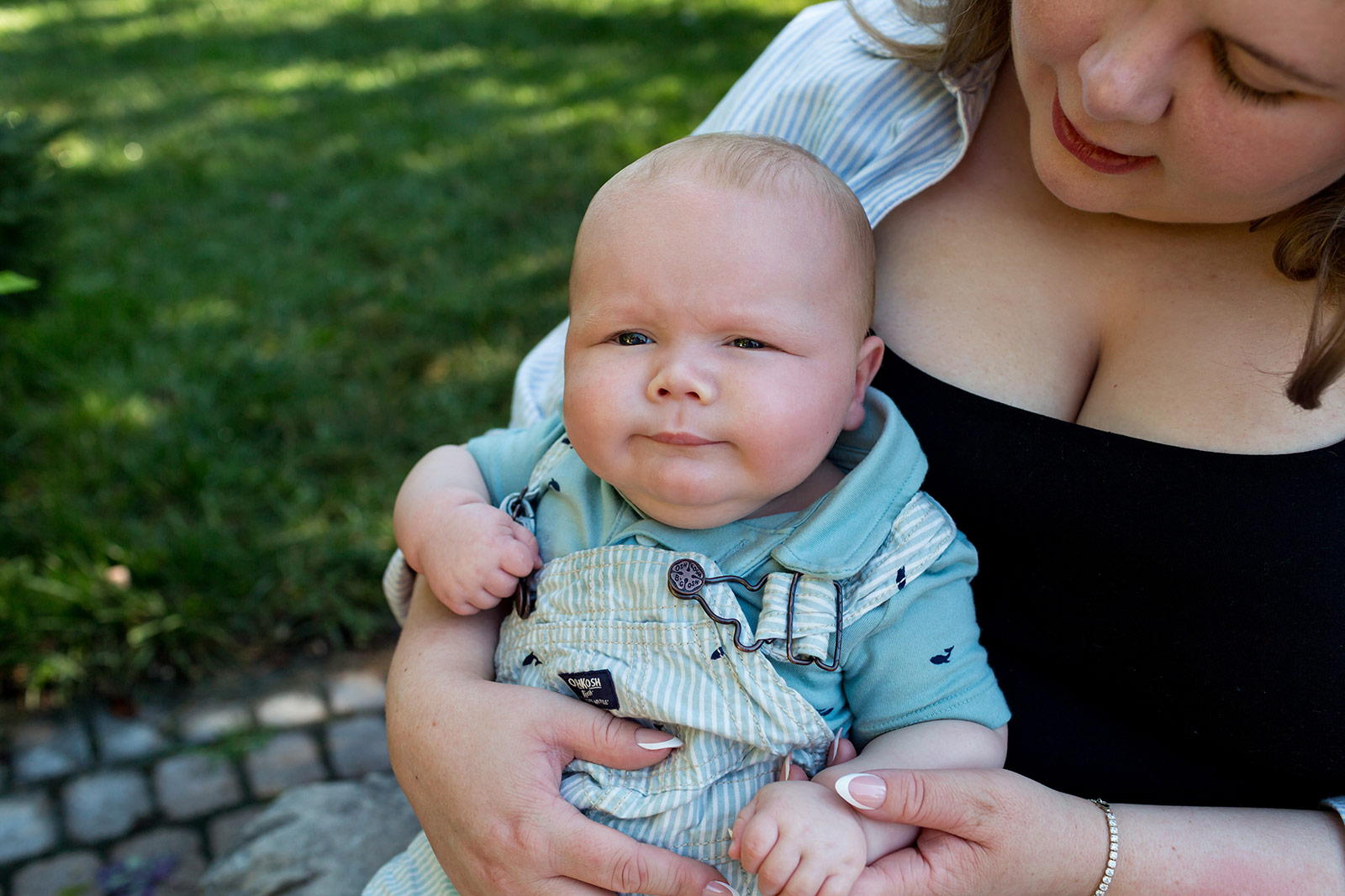 Close-up portrait of a baby in striped overalls sitting in mom's lap and looking curiously at the camera during an outdoor family session.